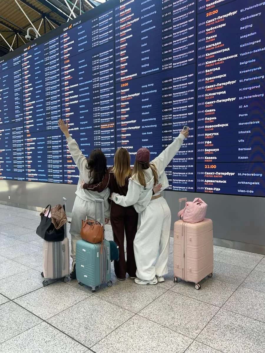 Three women with luggage standing in front of an airport departures board