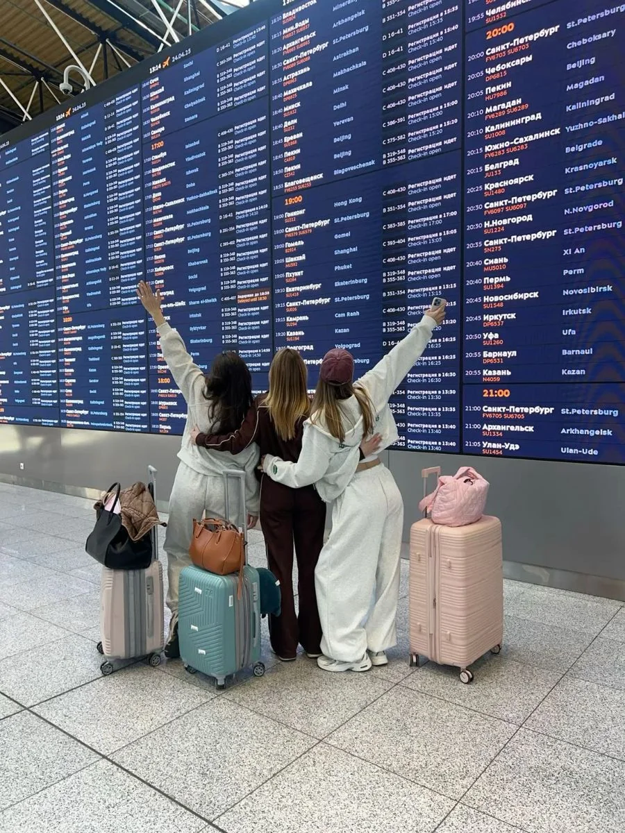 Three women with rolling luggage standing in front of an airport departures board